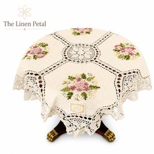 May include: Cream-colored, round tablecloth with embroidered floral bouquets in pink, green, and white. The tablecloth has a crocheted border and a central crocheted design. The Linen Petal logo is visible.