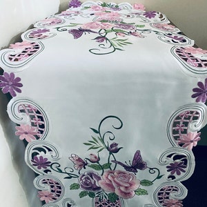 May include: White table runner with embroidered pink and purple flowers, butterflies, and leaves. The runner has a scalloped edge with a delicate lace design.