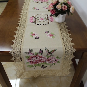 May include: A beige table runner with a lace border and embroidered floral and butterfly designs. The runner is on a wooden table with a vase of pink flowers.