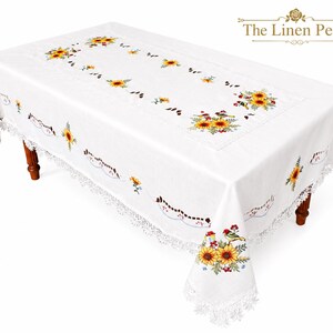 May include: White rectangular tablecloth with embroidered sunflowers and birds. The tablecloth has a lace trim and is set on a wooden table. The Linen Petal logo is in the upper right corner.