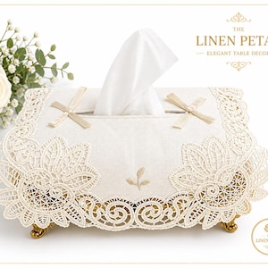 May include: An off-white tissue box cover with a cream-colored lace overlay and gold-colored feet. The cover features a floral lace design, cream-colored bows, and the text "THE LINEN PETAL - ELEGANT TABLE DECOR".