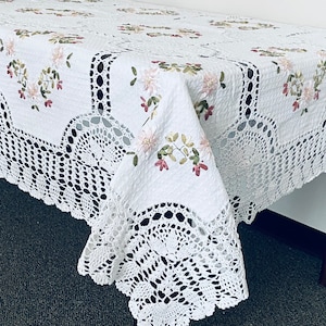 Pode incluir: Toalha de mesa de crochê branca com bordado floral delicado. A toalha de mesa tem uma borda festoneada e é feita de um tecido macio e leve.