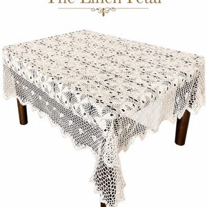May include: Cream-colored, rectangular crochet tablecloth with a scalloped edge. The tablecloth is draped over a dark brown wooden table. The text "The Linen Petal" is displayed at the top of the image.