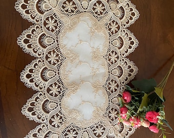 May include: Cream-colored lace table runner with a detailed floral design. The runner has a central panel and a scalloped edge. A bouquet of pink roses rests on the side, adding a touch of color to the scene.