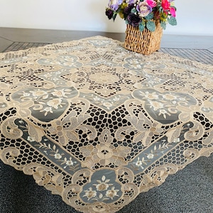 May include: A beige lace tablecloth with an intricate floral pattern. The tablecloth is square and has a delicate, embroidered design.