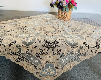 Puede incluir: Un mantel de encaje beige con un intrincado patrón floral. El mantel es cuadrado y tiene un delicado diseño bordado.