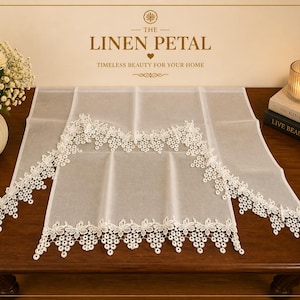 May include: A white linen curtain with intricate floral lace trim. The curtain is displayed on a dark wooden surface, with a vase of white roses and a lit candle in the background. The text "THE LINEN PETAL" is visible.