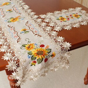 May include: Beige embroidered table runner with a sunflower design. The runner features yellow, orange, and green sunflowers with blue and red accents. The edges are trimmed with delicate, openwork lace.