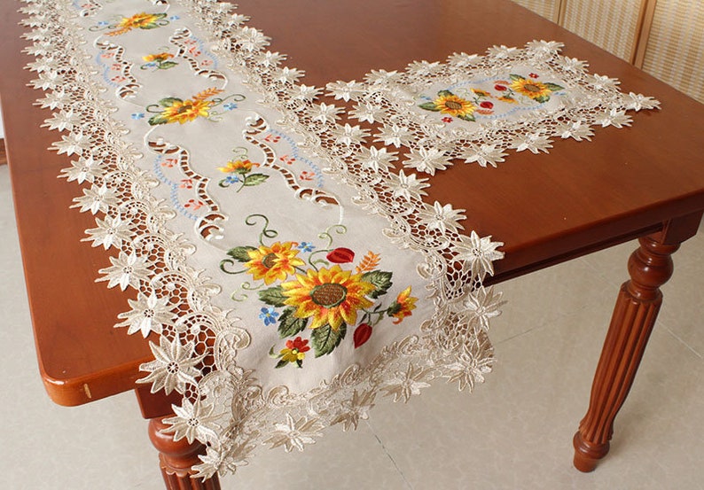 May include: Beige embroidered table runner with a sunflower design. The runner features yellow, orange, and green sunflowers with blue and red accents. The edges are trimmed with delicate, openwork lace.