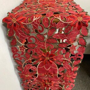 May include: Red and green embroidered table runner with a poinsettia design. The runner features a cut-out pattern and is perfect for adding a festive touch to your holiday table.