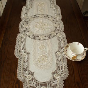 May include: Cream-colored lace table runner with intricate floral and leaf patterns. The runner features a central oval design and a scalloped edge. A teacup and saucer with a gold spoon are on the right side of the runner.