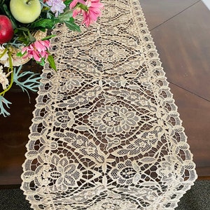 May include: A beige lace table runner with a delicate floral pattern. The runner is made of a fine, intricate lace and has a scalloped edge.