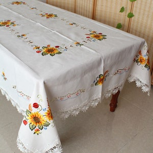 May include: White rectangular tablecloth featuring embroidered sunflowers and floral designs. The tablecloth has a delicate lace trim. The sunflowers are yellow, orange, and brown, with green leaves and stems.