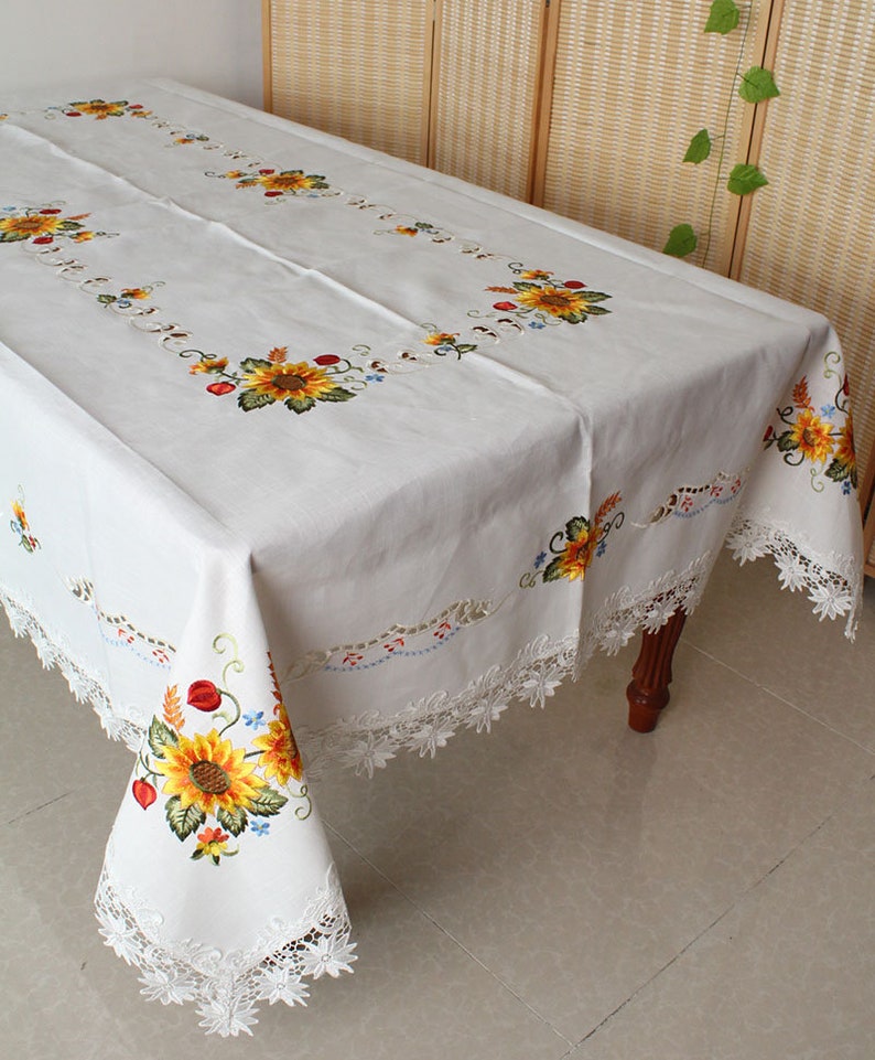 May include: White rectangular tablecloth featuring embroidered sunflowers and floral designs. The tablecloth has a delicate lace trim. The sunflowers are yellow, orange, and brown, with green leaves and stems.