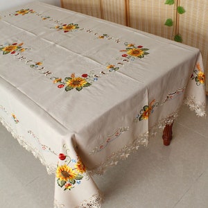 May include: Beige rectangular tablecloth with embroidered sunflowers, red berries, and blue flowers. The edges feature a delicate lace trim. The tablecloth is draped over a wooden table.