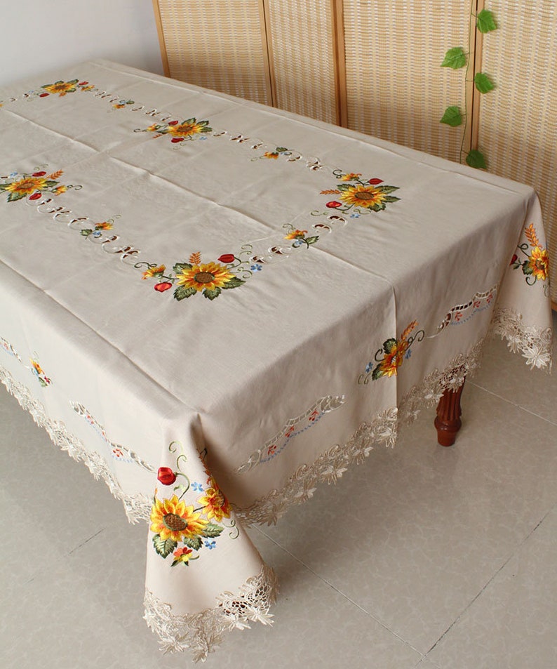 May include: Beige rectangular tablecloth with embroidered sunflowers, red berries, and blue flowers. The edges feature a delicate lace trim. The tablecloth is draped over a wooden table.