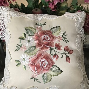 May include: Cream-colored square pillow with embroidered roses in shades of red and pink, surrounded by green leaves and small white flowers. The pillow has a delicate white lace trim around the edges, creating a vintage aesthetic.