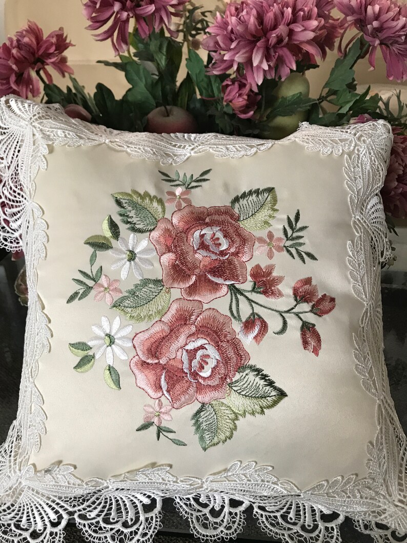 May include: Cream-colored square pillow with embroidered roses in shades of red and pink, surrounded by green leaves and small white flowers. The pillow has a delicate white lace trim around the edges, creating a vintage aesthetic.