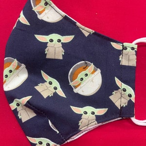 May include: A dark blue fabric face mask with a repeating pattern of green baby Yoda characters. Some of the characters are in a floating pod, while others are standing.