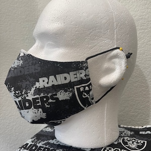 May include: Black and white face mask with a repeated pattern of the Las Vegas Raiders logo and the word "Raiders". The mask is on a white mannequin head.