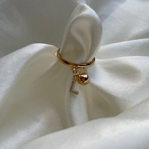 May include: A gold ring with a dangling charm featuring the letter L encrusted with clear rhinestones and a small gold ball.