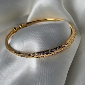 May include: A gold-toned open cuff bracelet featuring a textured surface with repeating star-shaped engravings. The bracelet has rounded, polished ball-shaped terminals at each end of the opening, providing a simple and clean design aesthetic.