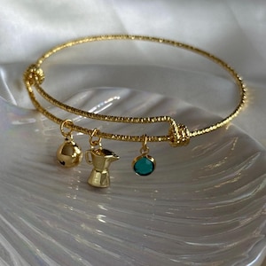 Puede incluir: Pulsera dorada con tres colgantes: una campana, una cafetera y una gema de color turquesa. La pulsera tiene un diseño texturizado y un detalle de nudo. Los colgantes cuelgan de la pulsera.