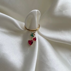Puede incluir: Un anillo dorado con un dije de cereza. El dije presenta dos gemas rojas en forma de corazón y detalles de hojas verdes. El anillo se exhibe sobre una tela blanca y sedosa.