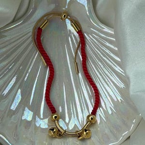 Può includere: Un braccialetto dorato con un design a cordoncino rosso. Il braccialetto presenta una barra dorata curva con tre piccole campane e dettagli dorati. Il cordoncino rosso è intrecciato e regolabile. Il braccialetto è esposto su un piatto bianco iridescente a forma di conchiglia.