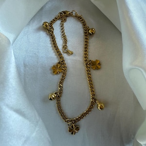 Puede incluir: Pulsera dorada con diseño de cadena de bordillo. Está adornada con abalorios de tréboles de cuatro hojas y pequeños colgantes en forma de campana. La joya se exhibe sobre una tela blanca y sedosa.