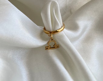 May include: A gold-colored ring with a Libra symbol charm. The ring is displayed on a white, silky fabric. The Libra symbol is a stylized balance scale, a common representation of the zodiac sign. The ring is a simple band with a small charm.