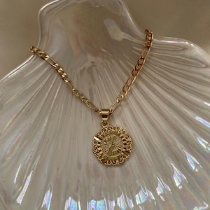 May include: A gold chain necklace with a round pendant featuring a stylized letter 'X' in the center. The necklace is displayed on a white seashell-shaped dish.