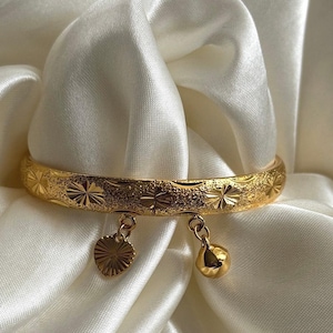 May include: A gold-coloured bangle bracelet with a textured surface and starburst and cross patterns. The bracelet has two dangling charms: a heart and a bell. The bracelet is displayed on white satin fabric.