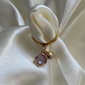 Puede incluir: Un anillo de oro con un dije de trébol morado y una pequeña campana dorada colgando.