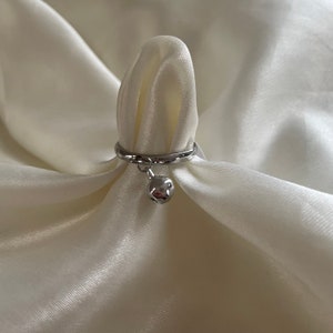 May include: A silver ring with a small silver bell charm hanging from it. The ring is displayed on a white fabric background.