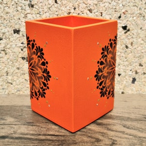 May include: Orange square-shaped pencil holder with black and gold mandala designs on each side.