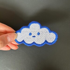 May include: Embroidered cloud patch with a smiling face, featuring a white centre and a blue outline. The patch is held against a dark background, showcasing its detailed stitching and cheerful design.