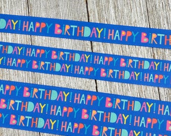 Könnte beinhalten: Drei blaue Bänder mit der Aufschrift "HAPPY BIRTHDAY!" in bunten Buchstaben. Die Bänder sind horizontal vor einem hellen Holzmaserungshintergrund angeordnet. Der Text ist in einem sich wiederholenden Muster.
