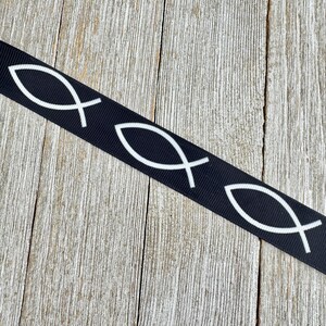 May include: Black ribbon featuring a repeating white Ichthys symbol, also known as the Jesus fish. The ribbon is set against a weathered, light brown wooden surface. The fish symbol is a Christian symbol.