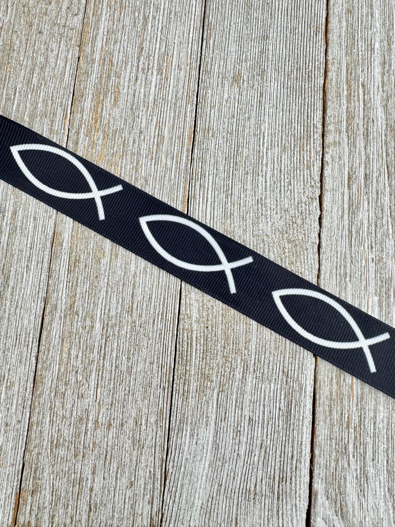 May include: Black ribbon featuring a repeating white Ichthys symbol, also known as the Jesus fish. The ribbon is set against a weathered, light brown wooden surface. The fish symbol is a Christian symbol.