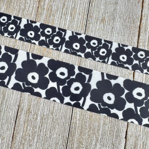 May include: Two ribbons with a black and white floral pattern. The flowers are stylized with rounded petals and a central white circle. The ribbons are on a light-colored wooden surface.