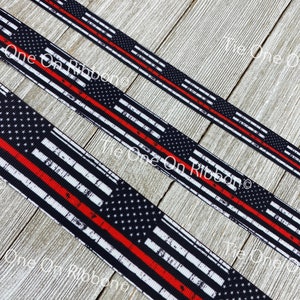 May include: Black and white distressed American flag ribbon with a single red stripe across the middle. The ribbon is on a wooden surface.