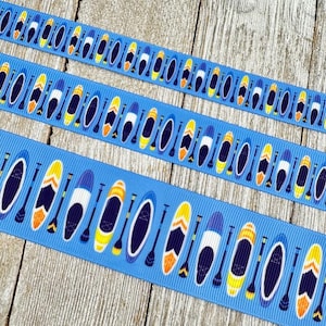 May include: Three strips of blue ribbon featuring a repeating pattern of stand-up paddleboards and paddles in various colours. The paddleboards are yellow, white, and blue. The ribbon is on a wooden surface.