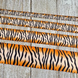 May include: Several ribbons with a tiger stripe pattern in orange, black, and cream are displayed against a weathered wood background. The ribbons vary in width, showcasing the repeating animal print design.