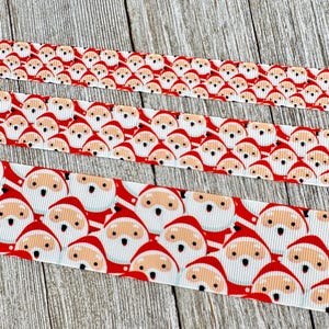 Peut inclure: Trois rubans avec un motif répété de visages du Père Noël. Les Pères Noël ont des bonnets rouges et des barbes blanches sur fond blanc. Les rubans sont rouges et blancs et sont placés sur une surface en bois.