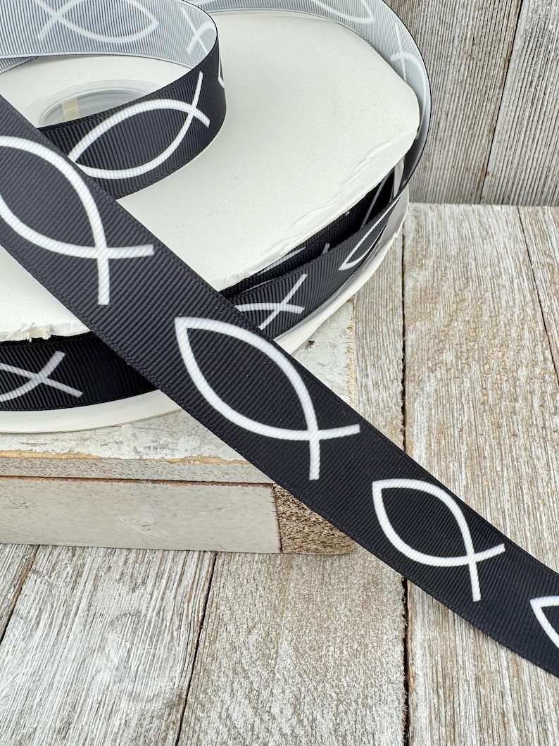 May include: Black ribbon with a repeating white fish symbol pattern. The ribbon is spooled on a white cardboard roll. The image is on a wooden surface.