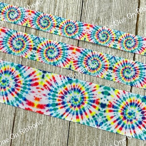 May include: Tie dye ribbon with a white background and colorful swirls of blue, red, yellow, and green. The ribbon is on a wooden surface.