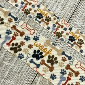 May include: White fabric ribbon with a repeating pattern of brown, blue, and tan paw prints, bones, and the word "woof" in yellow letters.