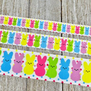 May include: Three ribbons with a repeating pattern of colorful bunny figures. The bunnies are in shades of blue, pink, yellow, and green, with black dot eyes. The ribbons are on a light wood grain background.