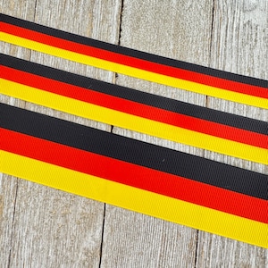 May include: Three ribbons with the colors of the German flag: black, red, and yellow. The ribbons are diagonally placed on a wooden surface. The colors are in horizontal stripes.